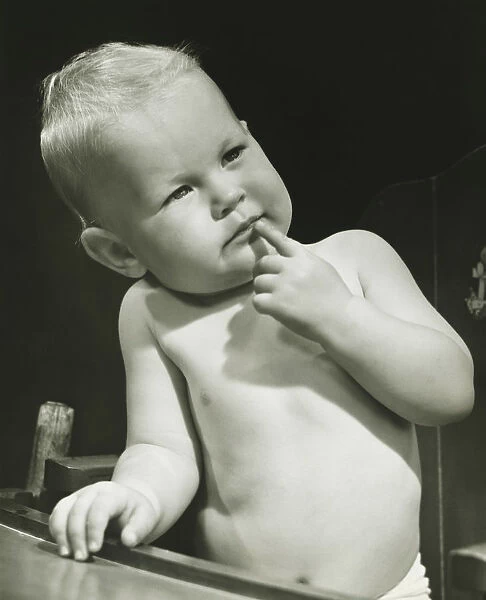 Baby boy (6-12 months) with finger on lips sitting in high chair, (B&W), portrait