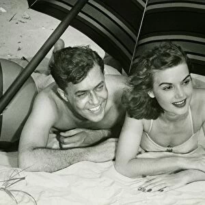 Young couple lying on sand under umbrella, smiling, (B&W)