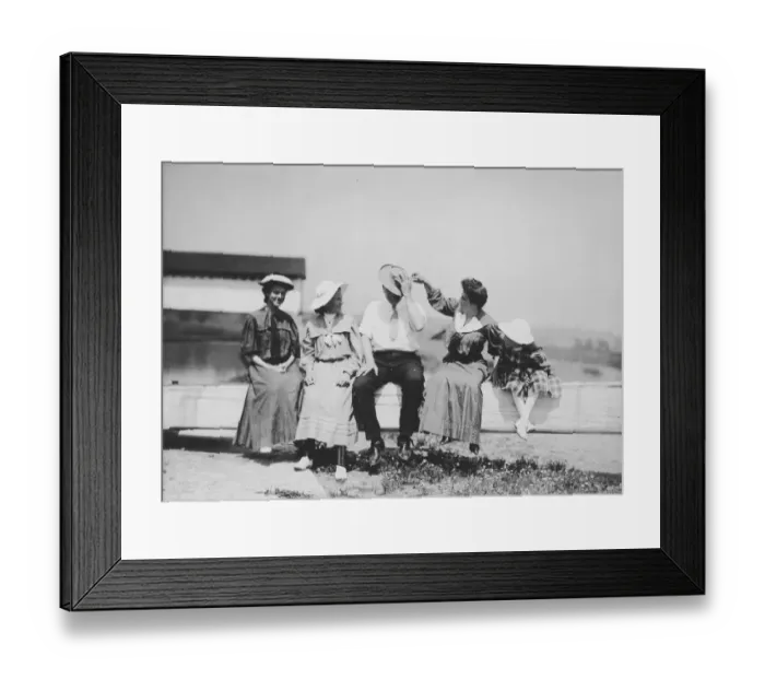 Framed Print : Fence Sitters; Three women, a man and a girl sit on a fence while one of the women Framed Print : Fence Sitters; Three women, a man and a girl sit on a fence while one of the women