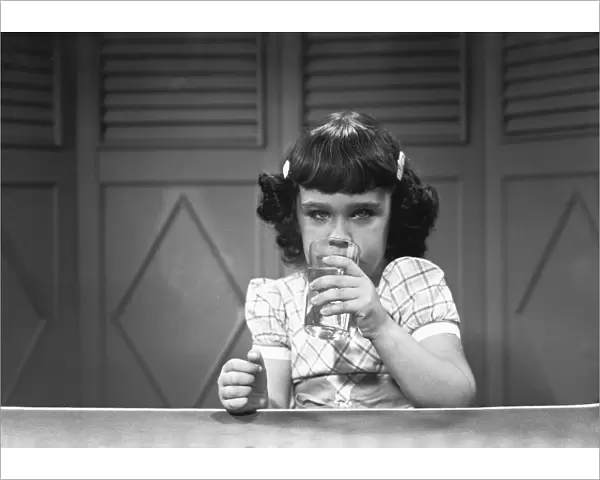 Girl (6-7) sitting at table, drinking water from glass, (B&W)