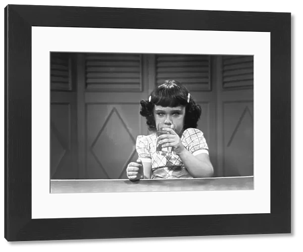 Girl (6-7) sitting at table, drinking water from glass, (B&W)