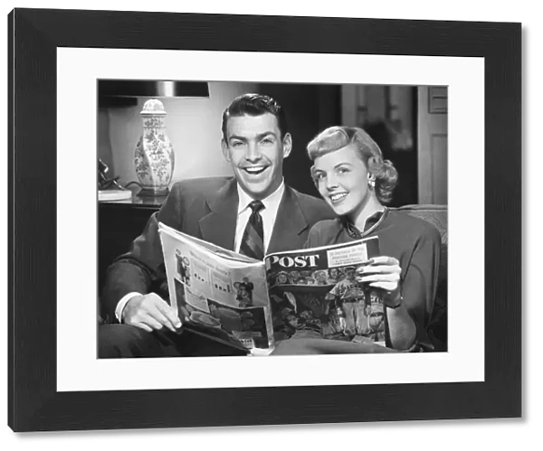 Couple sitting on sofa, holding magazine, (B&W), portrait