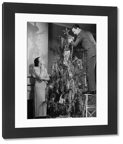 Woman assisting man placing star on top of Christmas tree, (B&W)