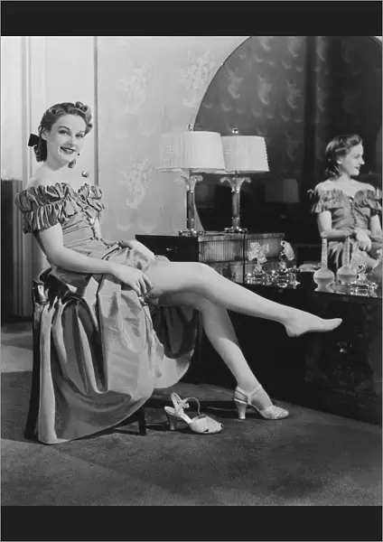 Woman sitting at vanity table, putting on stockings, (B&W), portrait