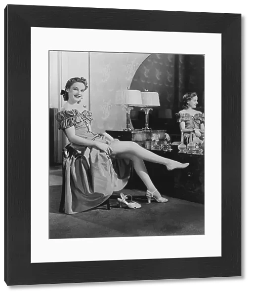 Woman sitting at vanity table, putting on stockings, (B&W), portrait