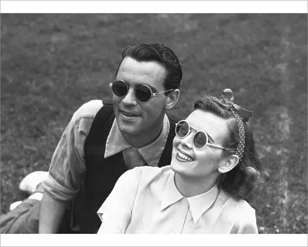 Couple wearing sunglasses sitting on grass, (B&W)