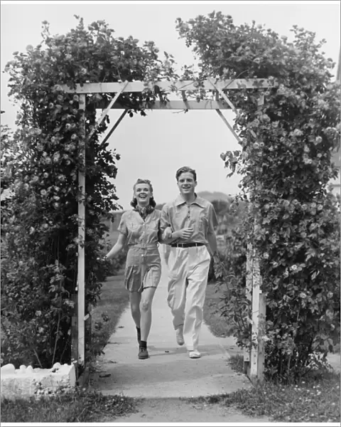 Couple walking on footpath towards rose covered pergola, (B&W)