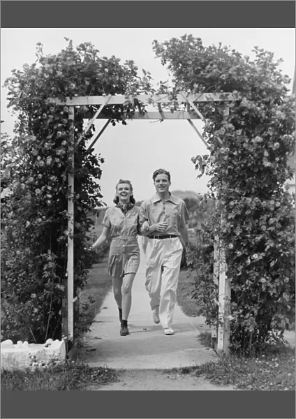 Couple walking on footpath towards rose covered pergola, (B&W)