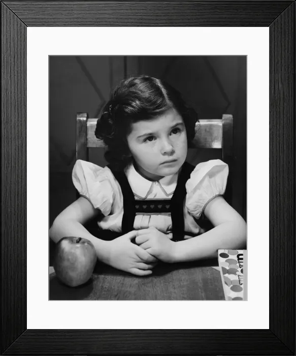 Framed Print : Girl (6-7) sitting at table, looking up, (B&W)