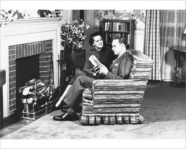 Couple sitting on armchair in front of fireplace, (B&W)