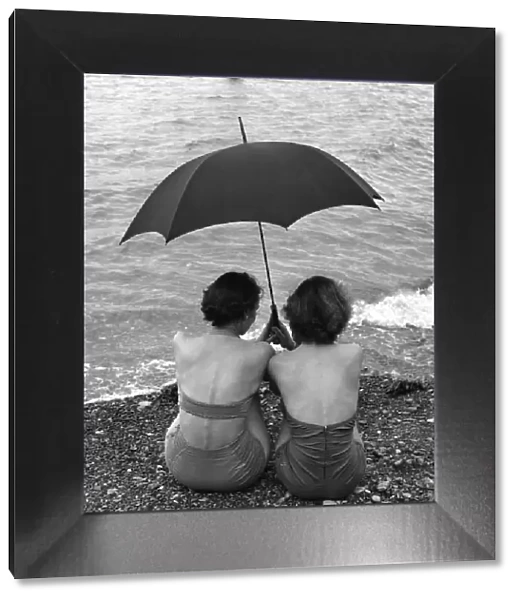 Rainy Holiday; Two women sheltering under their umbrella as they sit on the beach at Torquay in the rain