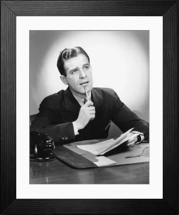 Framed Print : Man sitting at desk, holding pencil to lip, (B&W), portrait Framed Print : Man sitting at desk, holding pencil to lip, (B&W), portrait