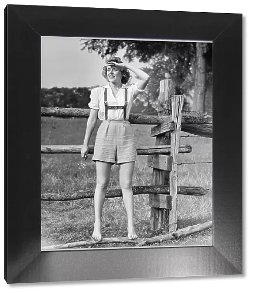 Barefoot woman in shorts standing on wooden fence on meadow, (B&W)