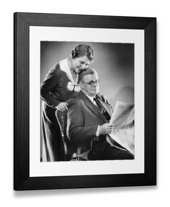 Framed Print : Couple at home, man reading newspaper, woman standing behind, (B&W)