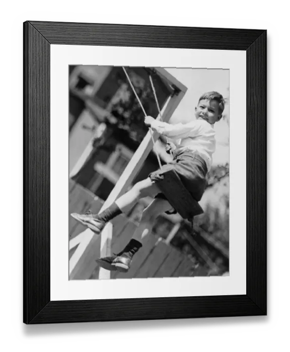 Framed Print : Boy (6-7) swinging in garden, (B&W), (Low angle view) Framed Print : Boy (6-7) swinging in garden, (B&W), (Low angle view)
