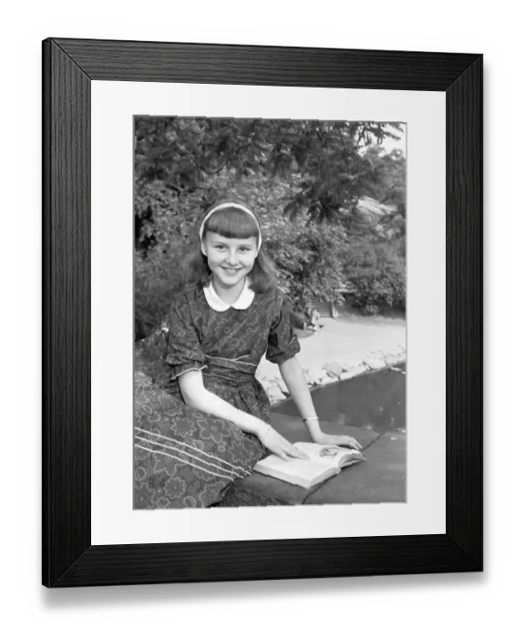 Framed Print : Girl (12-13) sitting on wall with book, (B&W), portrait