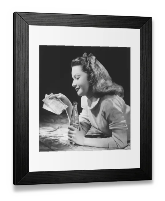 Framed Print : Teenage girl (16-17) lying on bed, reading letter, holding bottle (B&W) Framed Print : Teenage girl (16-17) lying on bed, reading letter, holding bottle (B&W)