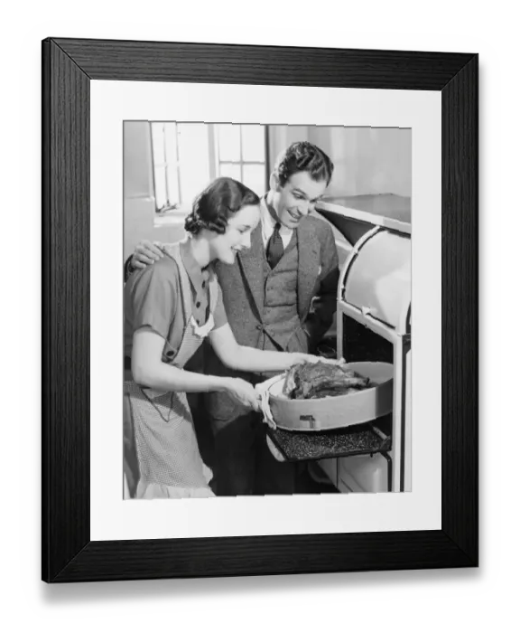 Framed Print : Couple in kitchen, wife taking roast from oven (B&W) Framed Print : Couple in kitchen, wife taking roast from oven (B&W)
