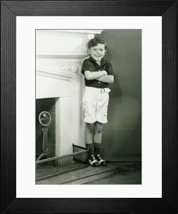 Framed Print : Young boy (6-7) standing by fireplace, (B&W) Framed Print : Young boy (6-7) standing by fireplace, (B&W)
