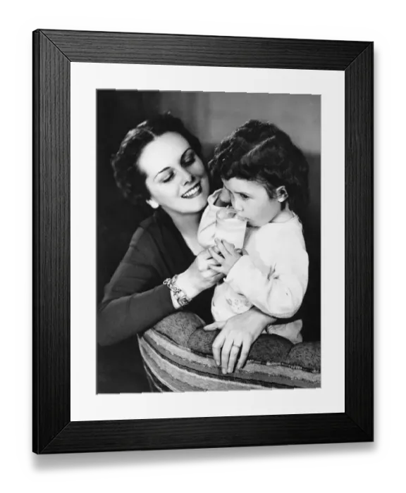 Framed Print : Mother on couch giving daughter (4-5) glass of milk, (B&W) Framed Print : Mother on couch giving daughter (4-5) glass of milk, (B&W)