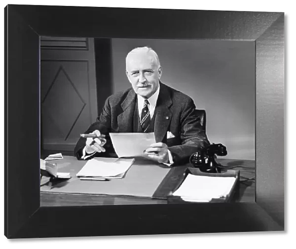 Senior businessman posing at desk in office (B&W), portrait