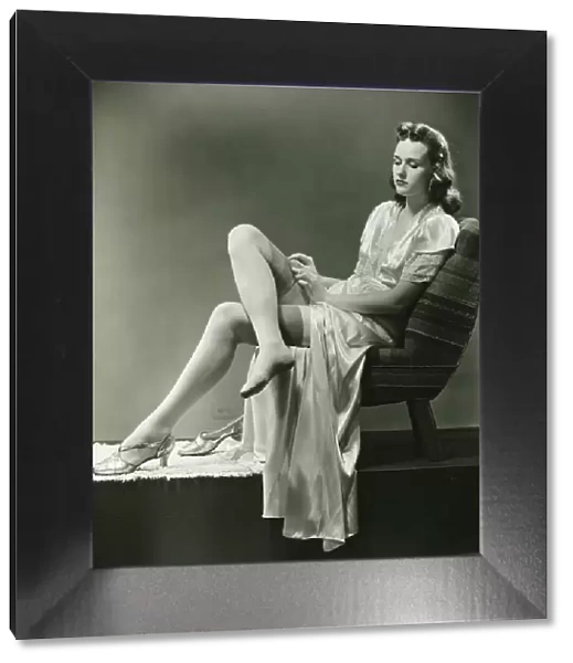 Young woman straightening stockings, sitting on chair in studio, (B&W)