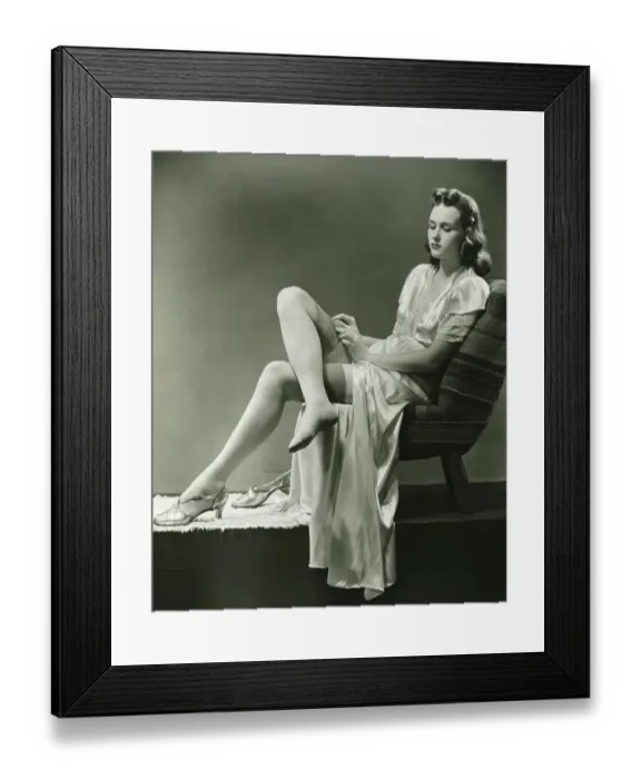 Framed Print : Young woman straightening stockings, sitting on chair in studio, (B&W) Framed Print : Young woman straightening stockings, sitting on chair in studio, (B&W)
