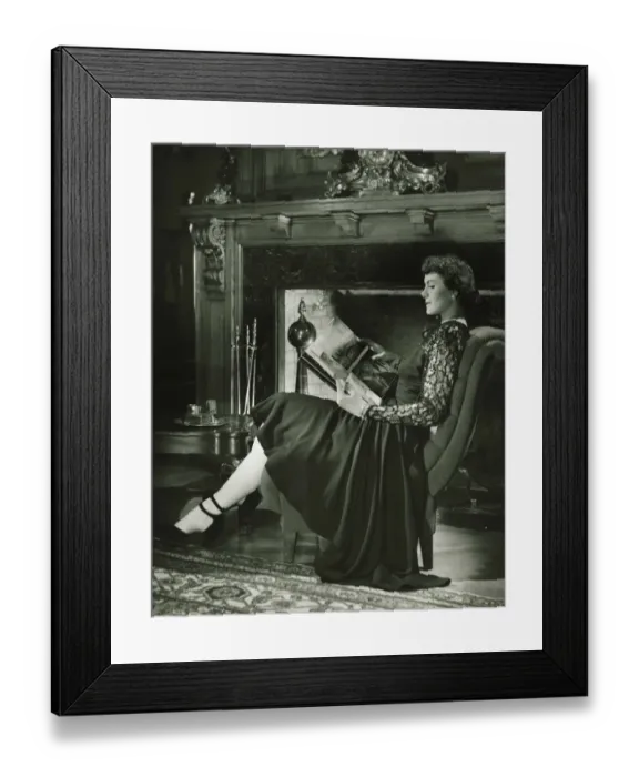 Framed Print : Young woman sitting by fireplace, holding album, (B&W) Framed Print : Young woman sitting by fireplace, holding album, (B&W)