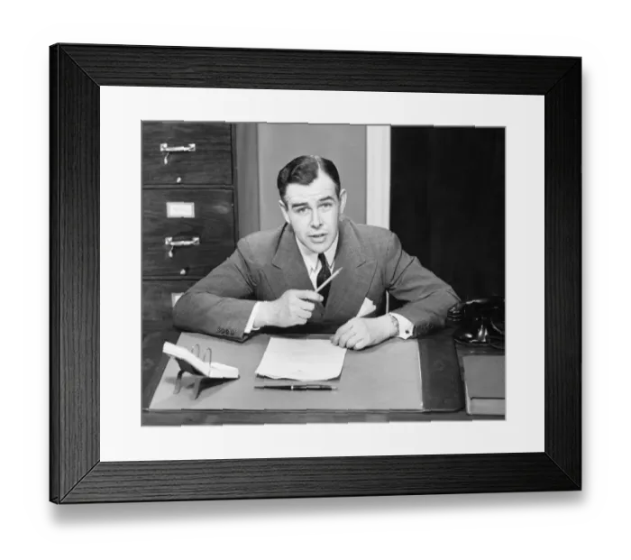 Framed Print : Young businessman sitting at small desk, holding pencil, (B&W)