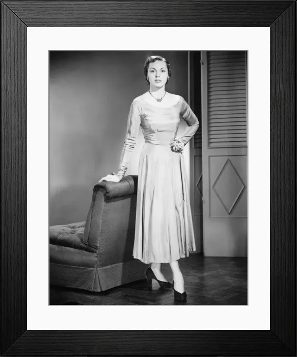 Framed Print : Young woman in evening dress standing, leaning against armchair, (B&W)