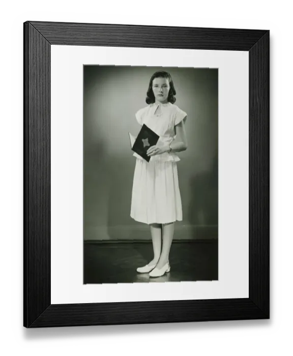 Framed Print : Teenage girl (13-14) in white dress standing in studio with open book, (B&W) Framed Print : Teenage girl (13-14) in white dress standing in studio with open book, (B&W)