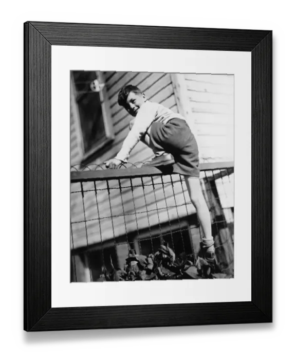 Framed Print : Boy (8-9) climbing on enclosure in front of house, (B&W) Framed Print : Boy (8-9) climbing on enclosure in front of house, (B&W)