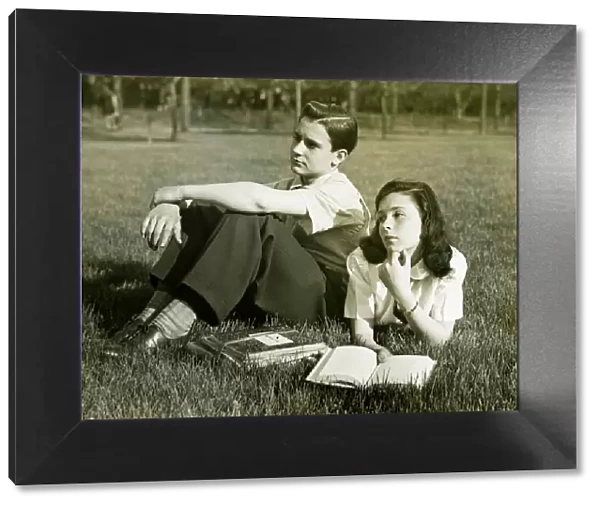 Two teenagers resting in field, boy (16-17) sitting, girl (14-15) lying with book, (B&W)