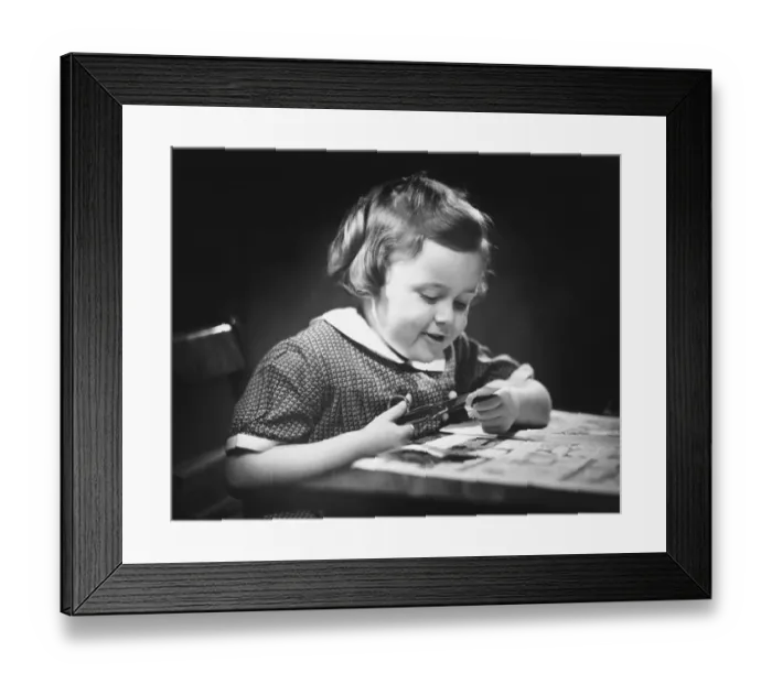Framed Print : Young girl (4-5) at table cutting paper with scissors, (B&W), close-up Framed Print : Young girl (4-5) at table cutting paper with scissors, (B&W), close-up