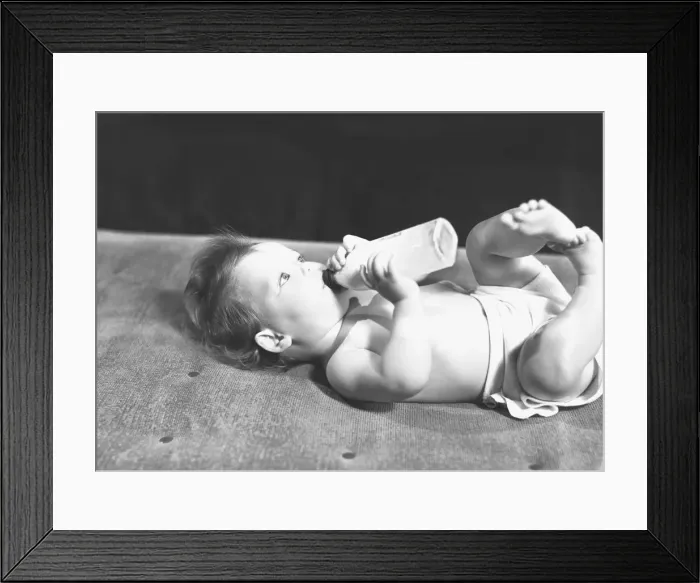 Framed Print : Baby (6-9 months) lying on bed, drinking from bottle, (B&W) Framed Print : Baby (6-9 months) lying on bed, drinking from bottle, (B&W)