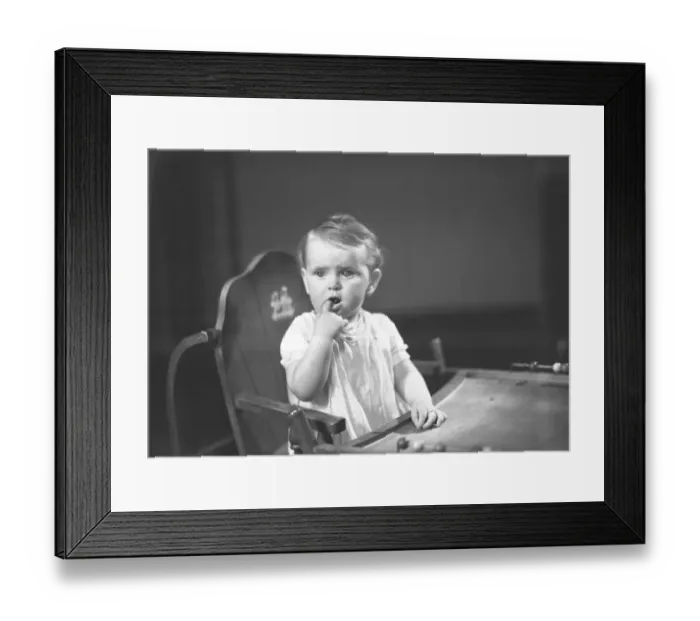 Framed Print : Baby girl (9-12 months) sitting in high chair, holding finger in mouth, (B&W)