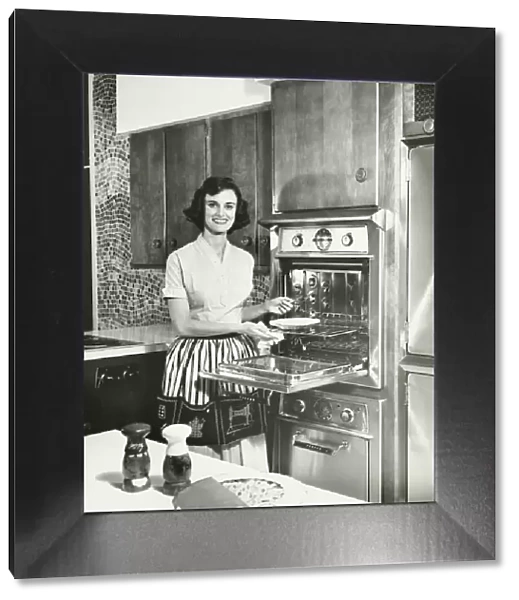 Woman putting pie into oven in kitchen, (B&W), portrait