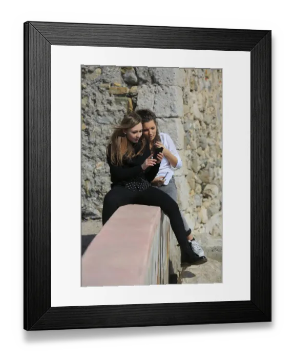Framed Print : Two girlfriends, teenagers, sitting on a wall and reading a text message on the mobile phone, Menton, Provence-Alpes-Cote dAzur, France Framed Print : Two girlfriends, teenagers, sitting on a wall and reading a text message on the mobile phone, Menton, Provence-Alpes-Cote dAzur, France