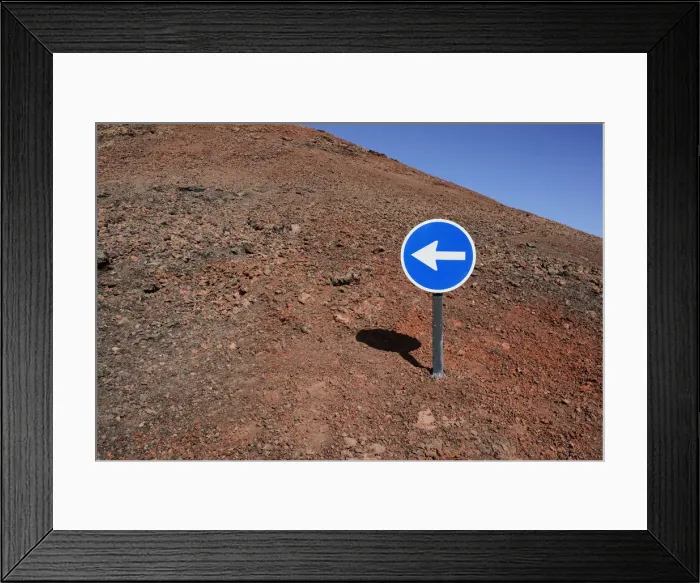 Framed Print : Blue sign with a white arrow pointing left, on red lava, Lanzarote, Canary Islands, Spain