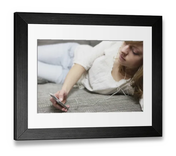 Framed Print : Young woman lying on a sofa with an MP3 player, listening to music through headphones Framed Print : Young woman lying on a sofa with an MP3 player, listening to music through headphones