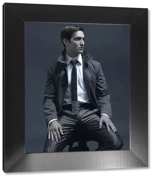 Fashion shot of a man in a suit and jacket, sitting on a stool