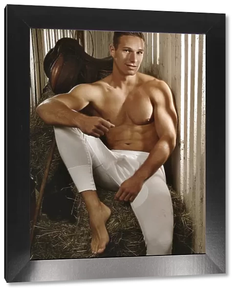 Man with a naked torso wearing long underwear in a horse barn sitting on bales of hay