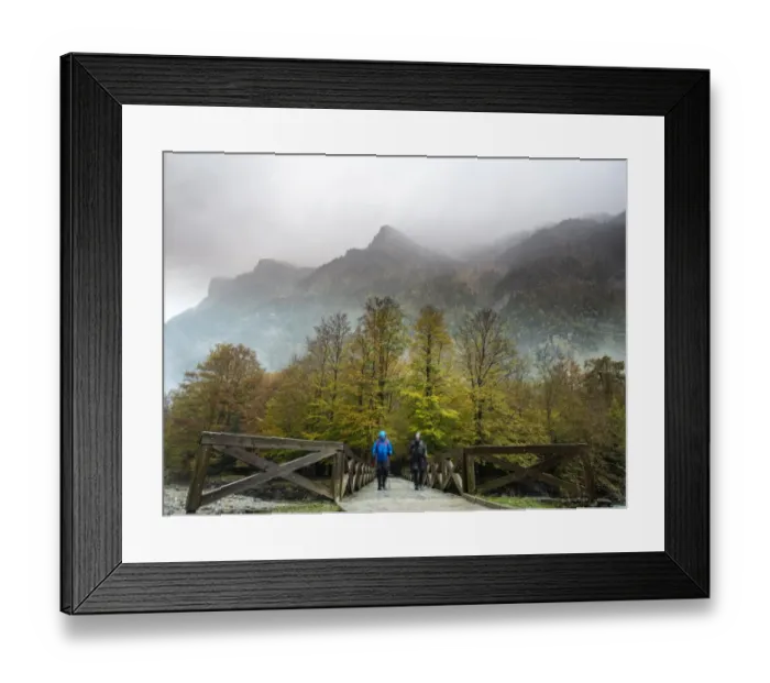 Framed Print : Hikers with raincoats and walking sticks on a bridge of wood that crosses a river in a forest in autumn