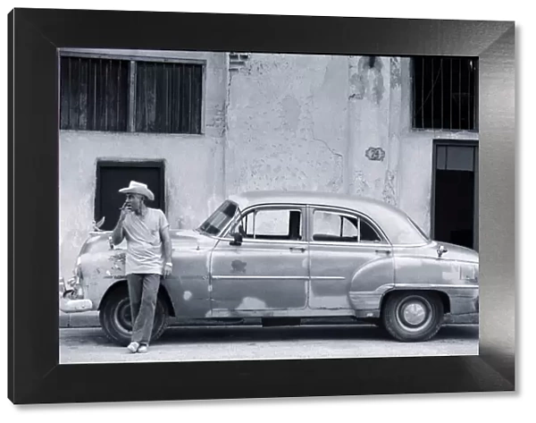 Cuban man leaning against car smoking cigar (B&W)
