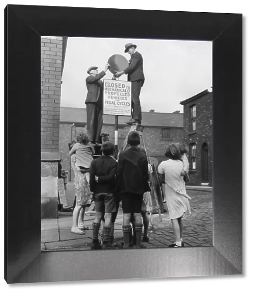 Play Safe. Children watch men putting up a sign closing their street to traffic