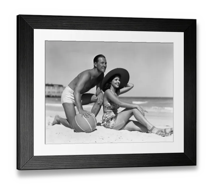 Framed Print : Couple on beach, man leaning on ball, woman sitting look out to ocean, wearing sun hat Framed Print : Couple on beach, man leaning on ball, woman sitting look out to ocean, wearing sun hat