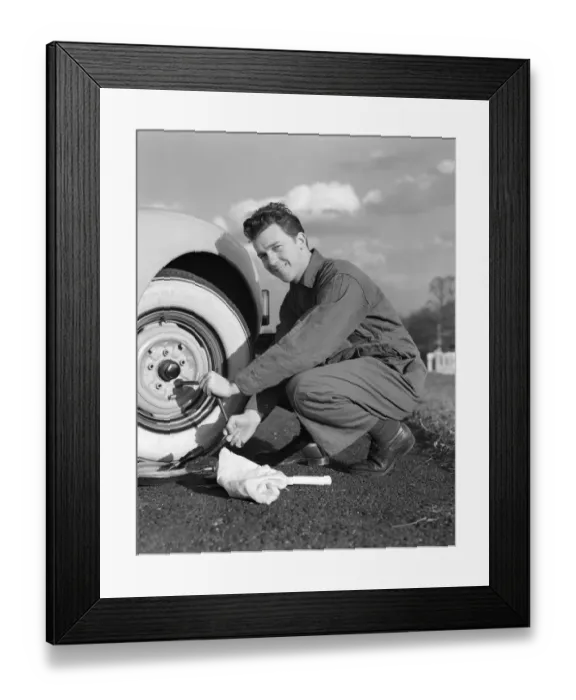 Framed Print : Smiling man in coveralls using tire iron to change tire on sedan