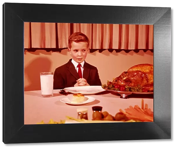 Boy sitting at table with hands folded for grace prayer, looking at roast turkey