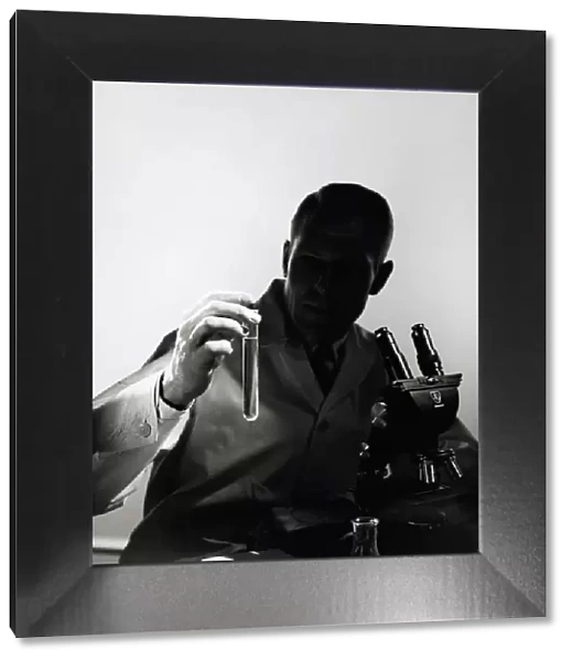 Male scientist wearing lab coat, sitting in darkness behind microscope