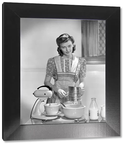Woman In A Apron Over A Cotton Print Dress Sifting Flour In A Bowl Between A Mixer & A